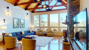 A spacious living room with a wooden ceiling, large windows, a stone fireplace, and a cozy seating area. - Angel Fire Country Club | Views | Camp Happy (Angel Fire)