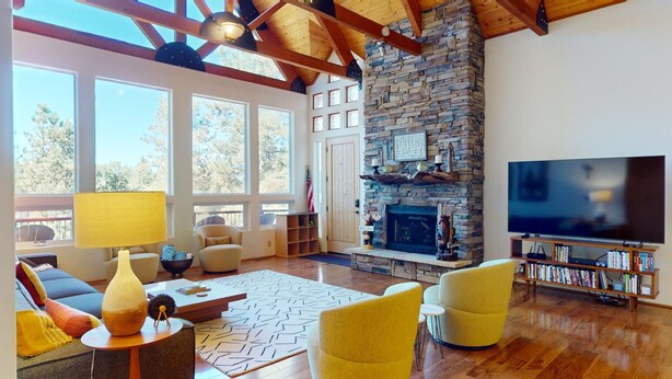 A spacious living room with a stone fireplace, large windows, a television, and a bookshelf. - Angel Fire Country Club | Views | Camp Happy (Angel Fire)