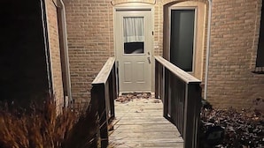 Une vue nocturne d’une maison en briques avec une terrasse en bois menant à une porte.