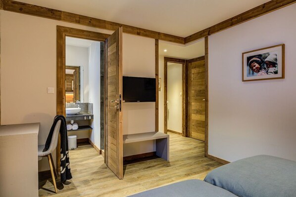 A room with a wooden ceiling, a television mounted on the wall, a bed, a chair, and a small table with a sink and mirror.