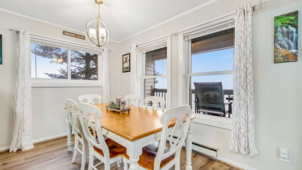 Un coin repas avec une table en bois et des chaises blanches, de grandes fenêtres avec des rideaux et un lustre.