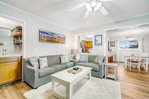 Un salon moderne avec un canapé d’angle gris, une table basse blanche et un coin repas comprenant une table et des chaises blanches.