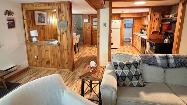 Ein gemütliches Wohnzimmer mit Sofa und Sessel, Holzwänden, einer Küchenzeile und einer Treppe, die nach oben führt.