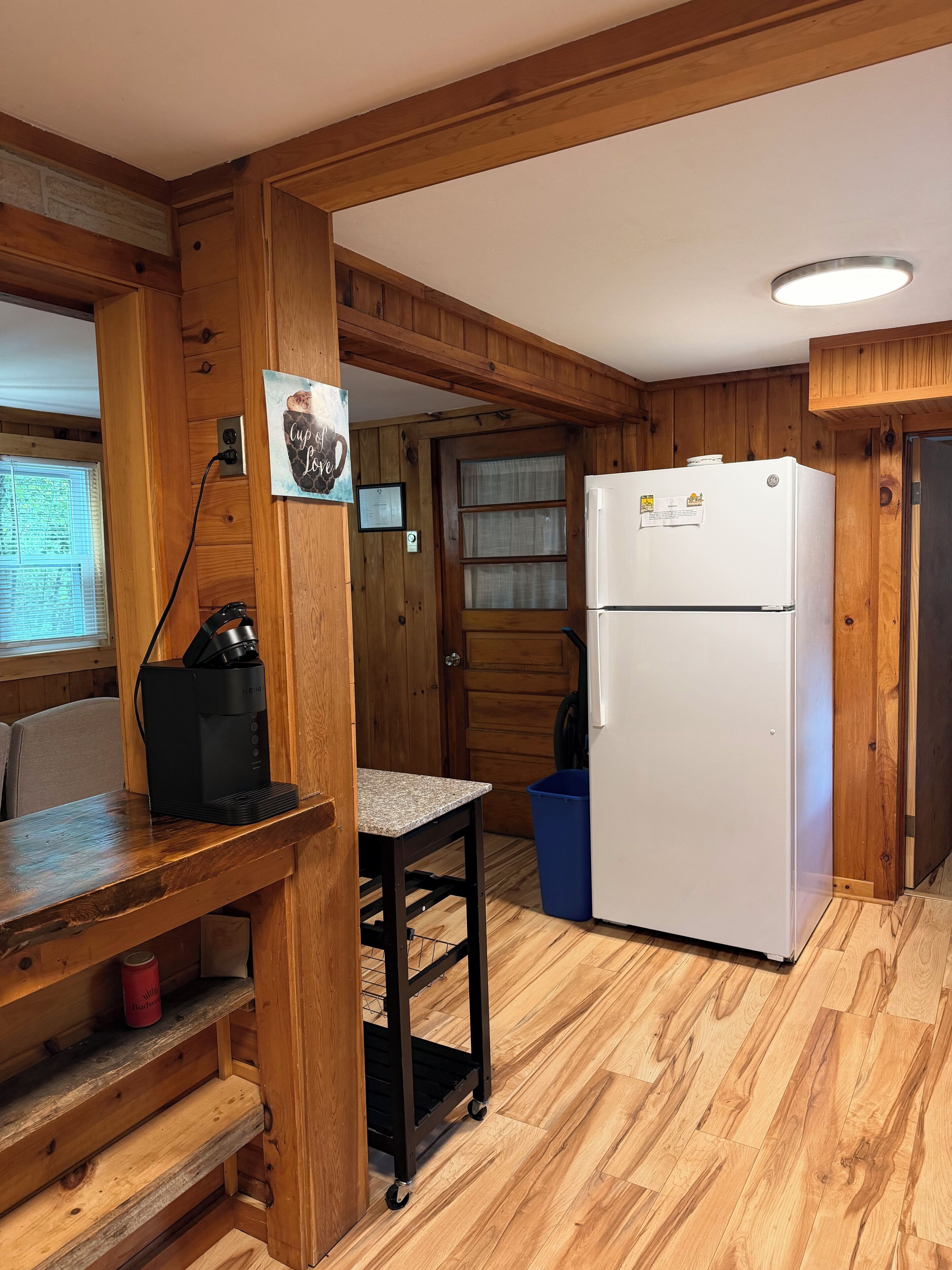 A compact kitchen with wooden walls, a white refrigerator, a coffee maker, and a window with blinds.