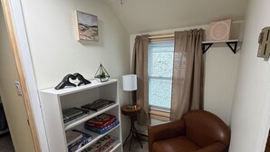 Ein Zimmer mit Bücherregal, braunem Ledersessel, kleinem Tisch mit Lampe und einem fenster mit Vorhängen.