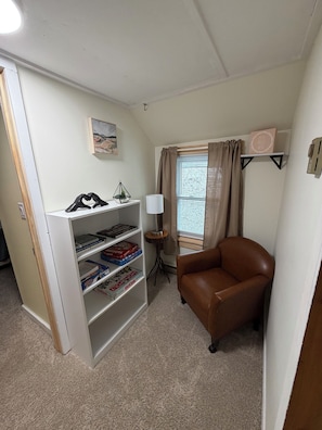 Una habitación con una estantería, un sillón de cuero marrón, una mesita con lámpara y una ventana con cortinas.
