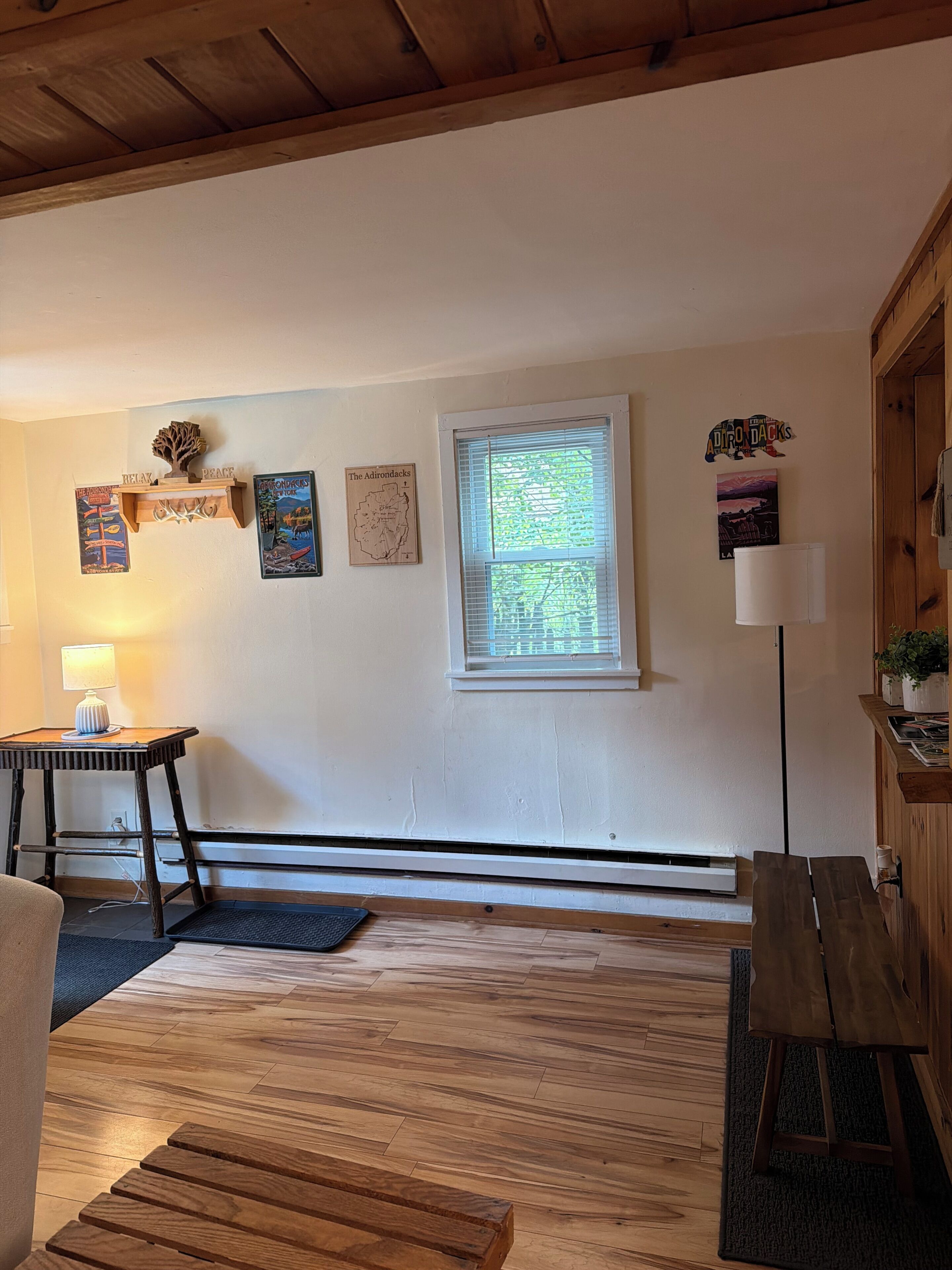 Una habitación con piso de madera, un escritorio con lámpara, una ventana con persianas, y adornos de pared que incluyen un mapa y un cartel.