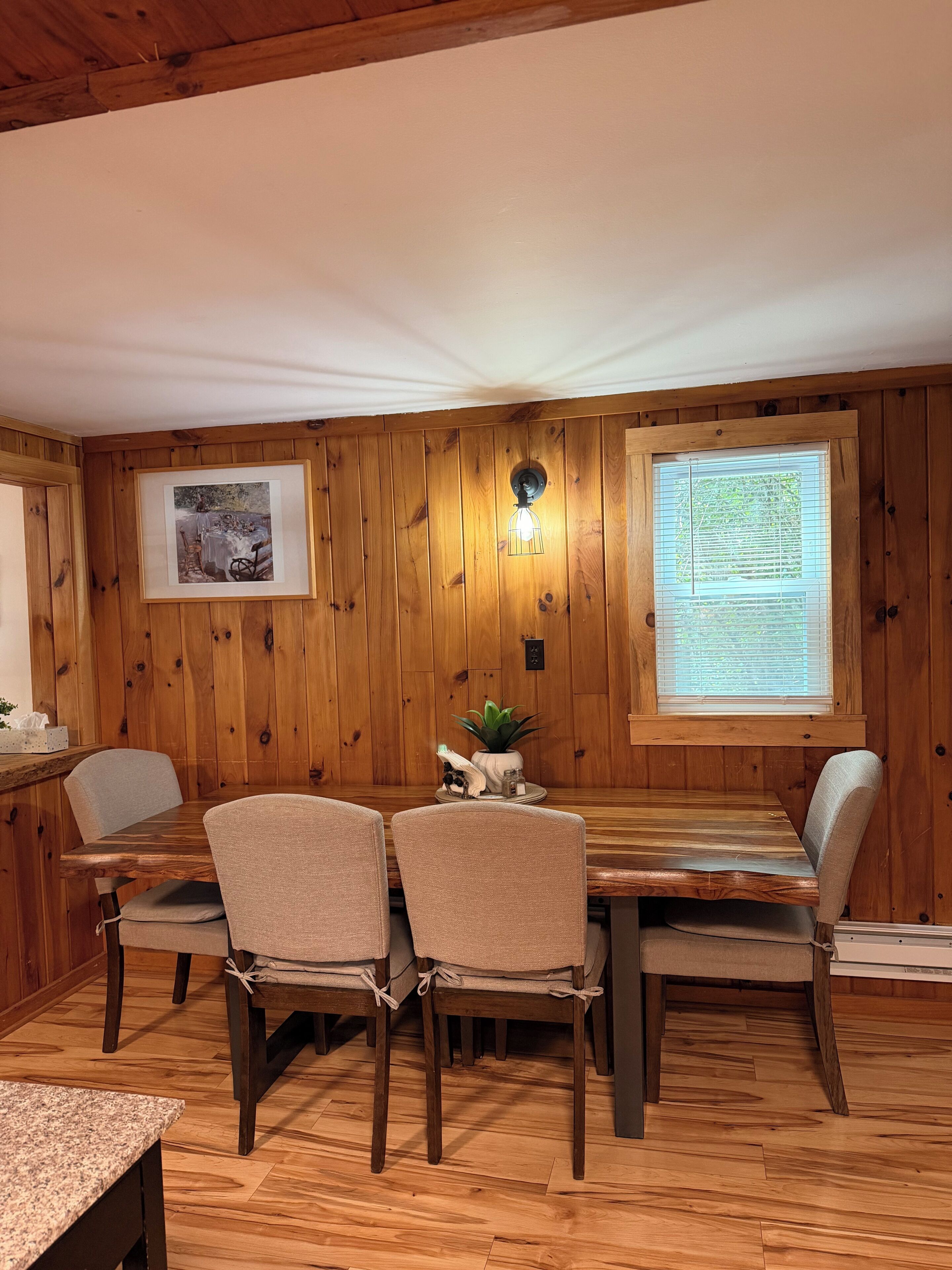 A wooden-paneled room with a dining table, chairs, a framed picture, a wall-mounted light fixture, and a window with blinds.