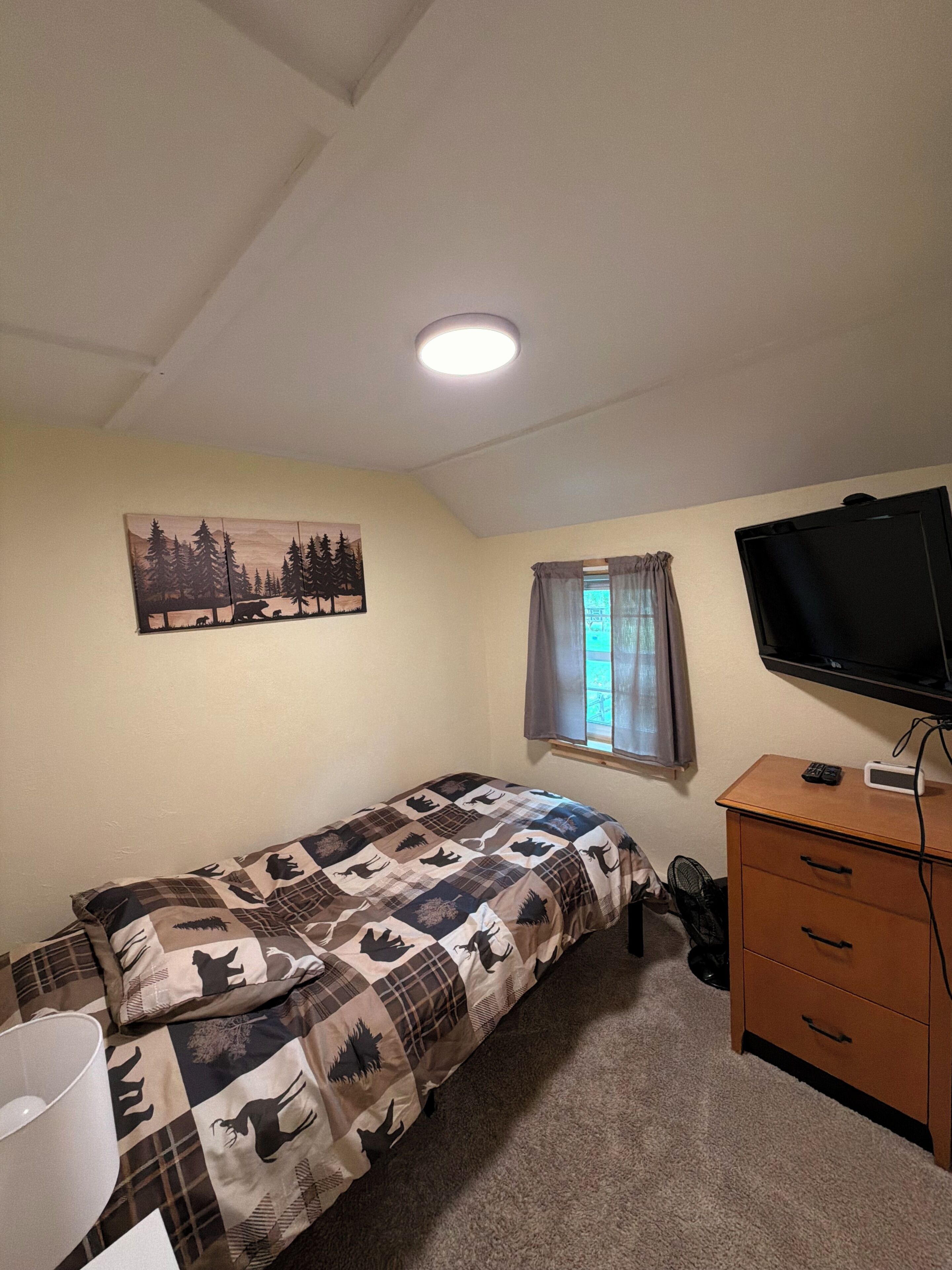 Una habitación pequeña y sencilla con cama, televisión, cómoda y ventana con cortinas.