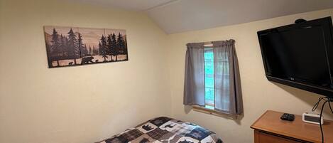 Una habitación pequeña y sencilla con cama, televisión, cómoda y ventana con cortinas.