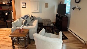 Ein Wohnzimmer mit einer Couch, einem Sessel, einem Couchtisch, Holzboden, einer Küchenzeile mit Geräten und einem Kamin.