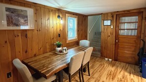 A wooden-paneled room with a dining table, chairs, a framed picture, a window with blinds, and a door. - The Cozy Cottage (Lake Placid)