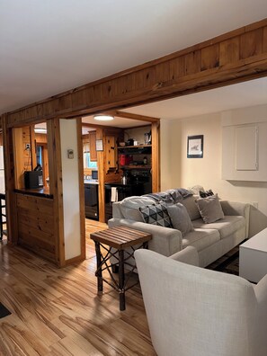 A living room with a wooden ceiling, a sofa, a coffee table, and a kitchen in the background. - The Cozy Cottage (Lake Placid)
