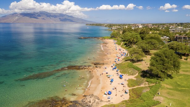 Una playa con aguas cristalinas, una montaña al fondo y un camino que lleva a un edificio.