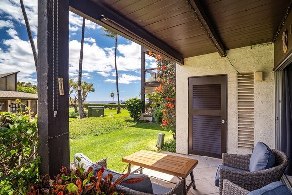 A covered patio with a table and wicker chairs, overlooking a well-maintained lawn and a view of the ocean.