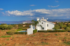 A white house with a pool, surrounded by dry vegetation and mountains in the background.