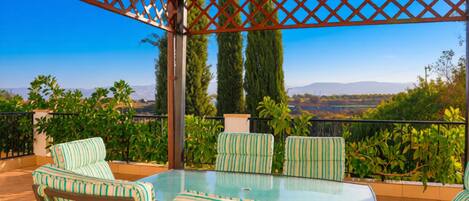 An outdoor patio with a glass table and striped chairs, surrounded by a wooden pergola and a view of greenery and mountains.