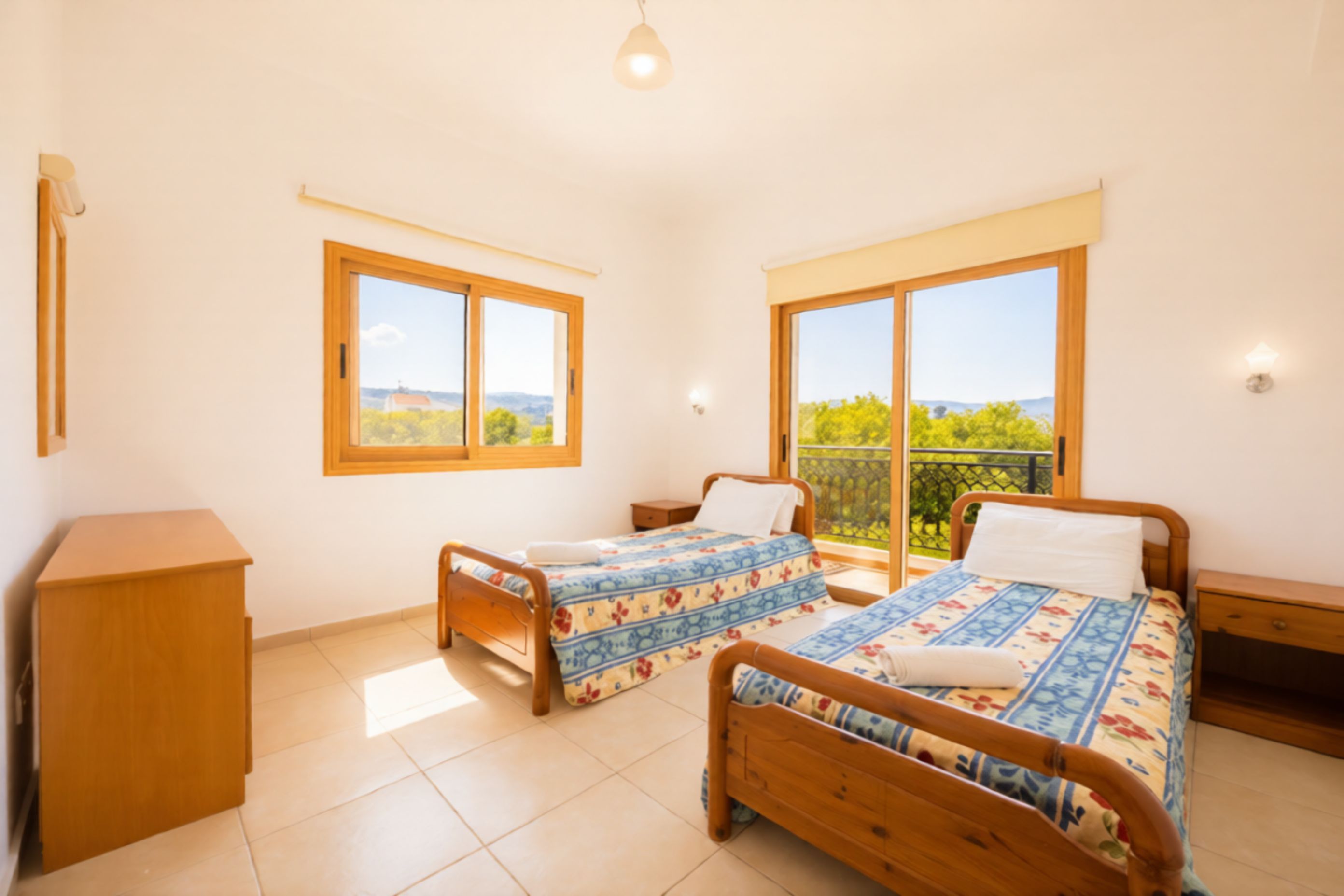 A bedroom with two single beds, a wooden dresser, and large windows with a view of greenery.