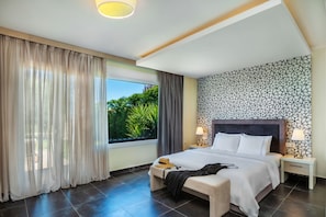 A modern hotel room with a large bed, a bedside table with lamps, a window with sheer curtains, and a patterned wall.