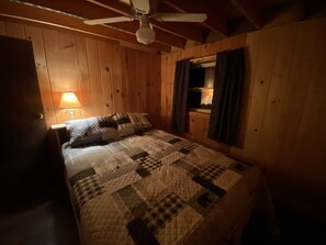 Un dormitorio de cabaña de madera con una cama, un ventilador de techo y una ventana con cortinas.