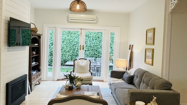 A living room with a fireplace, a sofa, a coffee table, and a dining area. - When a little white house in the country is all that is required! (Maffra)