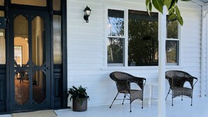 A white house with a black front door, two windows, a porch with wicker furniture, and a potted plant. - When a little white house in the country is all that is required! (Maffra)