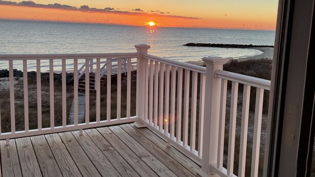 Eine Holzterrasse mit weißen Geländern mit Blick auf einen Strand bei Sonnenuntergang.