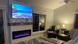 Ein modernes Wohnzimmer mit Flachbildfernseher über einem Kamin, Deckenventilator und großem Fenster mit Jalousien.
