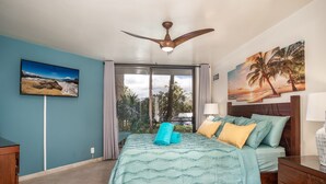 Ein Schlafzimmer mit einem Bett, einem Deckenventilator, einem Fenster mit Vorhängen und einem Bild von einer Strandlandschaft.