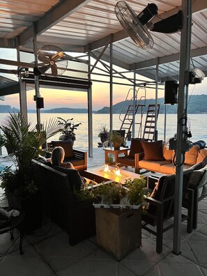 Ein Außenbereich zum Sitzen mit einem Feuerbereich, Ventilatoren und Blick auf das Wasser.