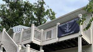 A two-story building with a staircase leading to a balcony. A banner reads "ARRR! YOU HERE YET?" and "PIRATE'S DEN". - BEST dock on lake.  New Hot Tub 2024. 4 BDRM, SLEEP 15.Aargh You Here Yet? (Gravois Mills)