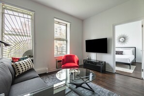 A modern living room with a sofa, a red armchair, a glass coffee table, a flat-screen TV, and a bed with a white headboard.