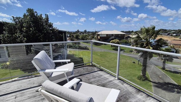 A balcony with two white lounge chairs, a glass balustrade, and a view of a residential area with houses and palm trees. - Sun trap with ocean views (Mangawhai Heads)