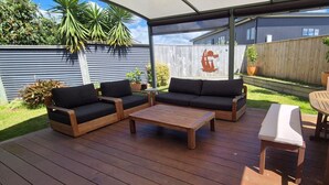 An outdoor patio with a wooden deck, two black sofas, a wooden coffee table, and a white table. - Sun trap with ocean views (Mangawhai Heads)