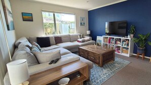 A living room with a sofa, coffee table, TV, bookshelf, and a potted plant. - Sun trap with ocean views (Mangawhai Heads)