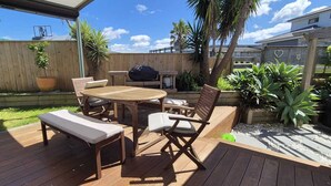 A wooden deck with a round table and chairs, a bench, and potted plants. - Sun trap with ocean views (Mangawhai Heads)