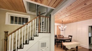 Un escalier en bois, doté de rampes blanches, mène à une salle à manger agrémentée d’un lustre.