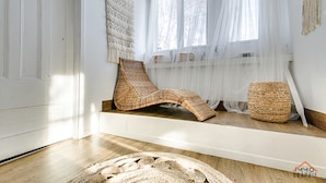 Um quarto com piso de madeira, uma cadeira de fibra natural, um banquinho de fibra natural e um tapete grande de fibra natural.