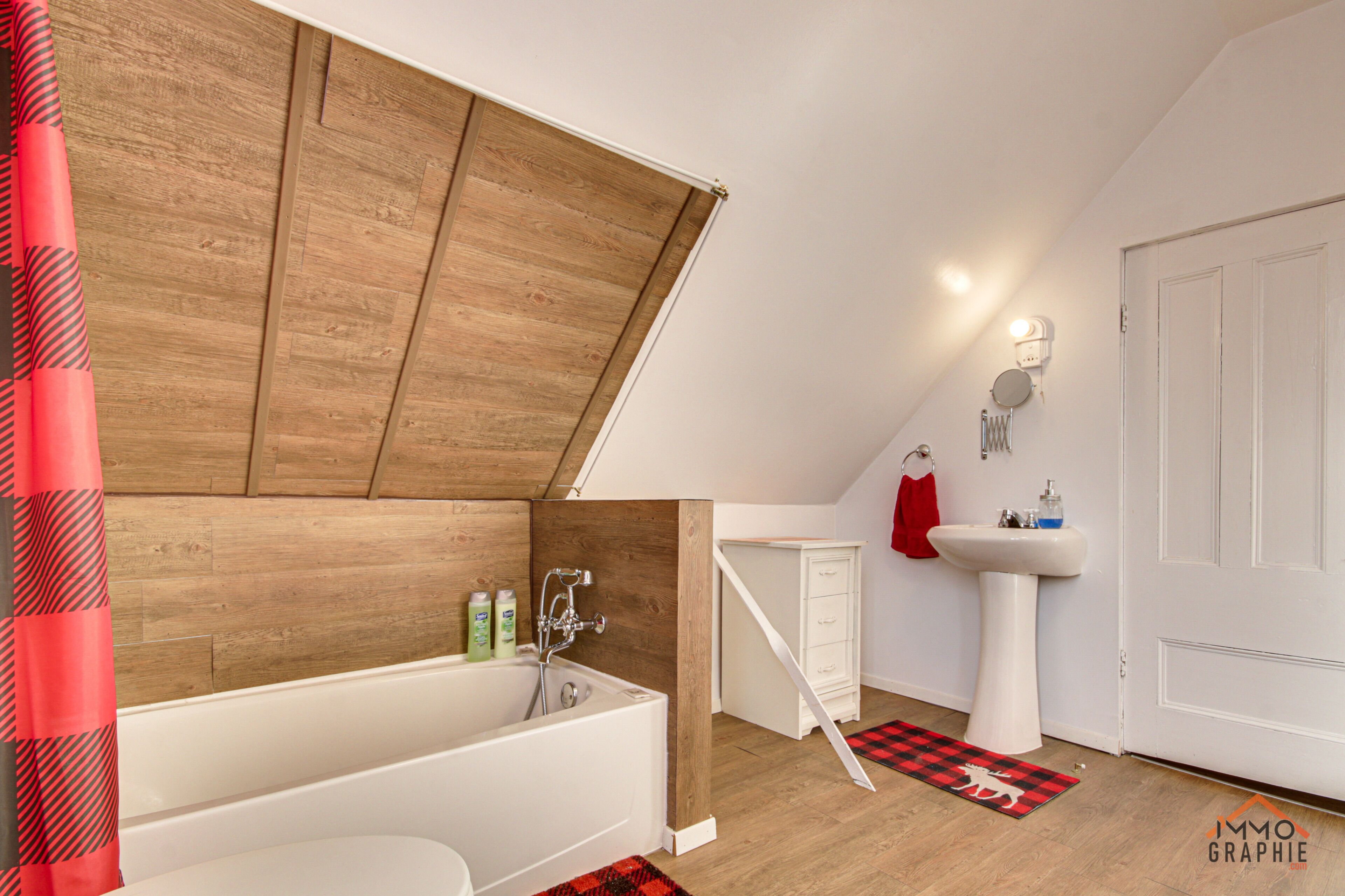 Une salle de bain moderne avec un plafond en pente en bois, une baignoire blanche, un lavabo blanc et un tapis à carreaux rouges et noirs.
