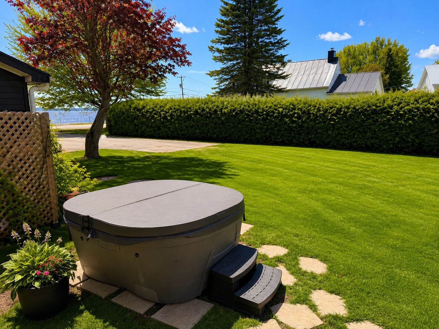 Un bain à remous dans un jardin bien entretenu, avec une pelouse, une haie et un bâtiment en arrière-plan.