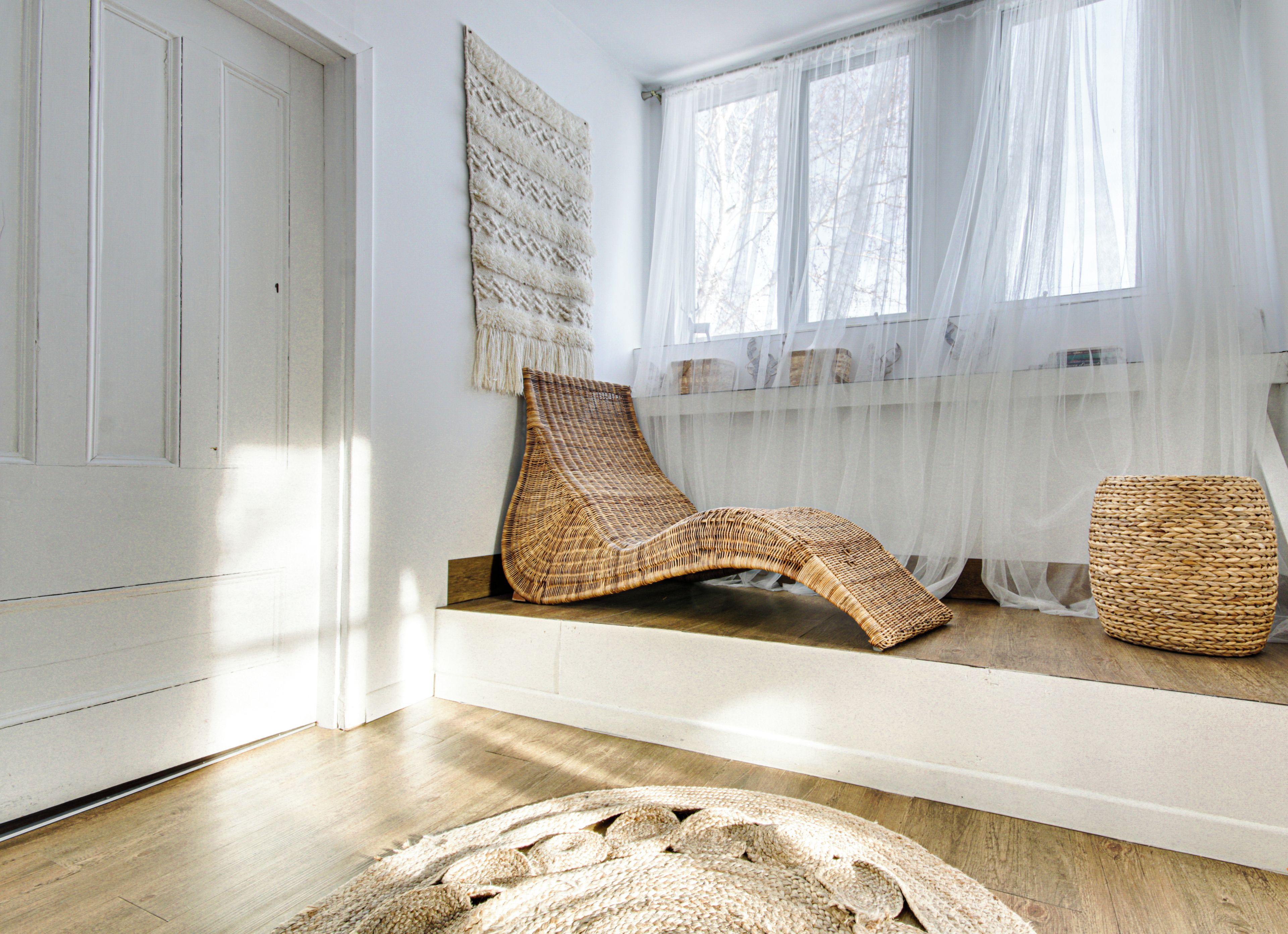 Una habitación con piso de madera, una silla tejida, un taburete tejido y un tapiz grande tejido.