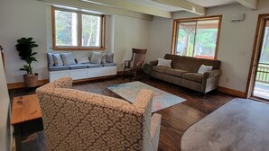 A spacious living room with a high ceiling, wooden beams, a sofa, a chair, and a large window providing natural light. - The Classic Cottage - Only one mile from the dunes! (Mears)