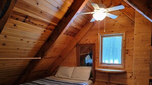 A cozy attic bedroom with a sloped ceiling, a bed with striped bedding, a ceiling fan, and a window with a view of trees. - The Classic Cottage - Only one mile from the dunes! (Mears)