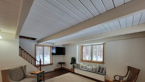 A spacious living room with a high ceiling, a wooden staircase, a flat-screen TV, and a bench with cushions. - The Classic Cottage - Only one mile from the dunes! (Mears)