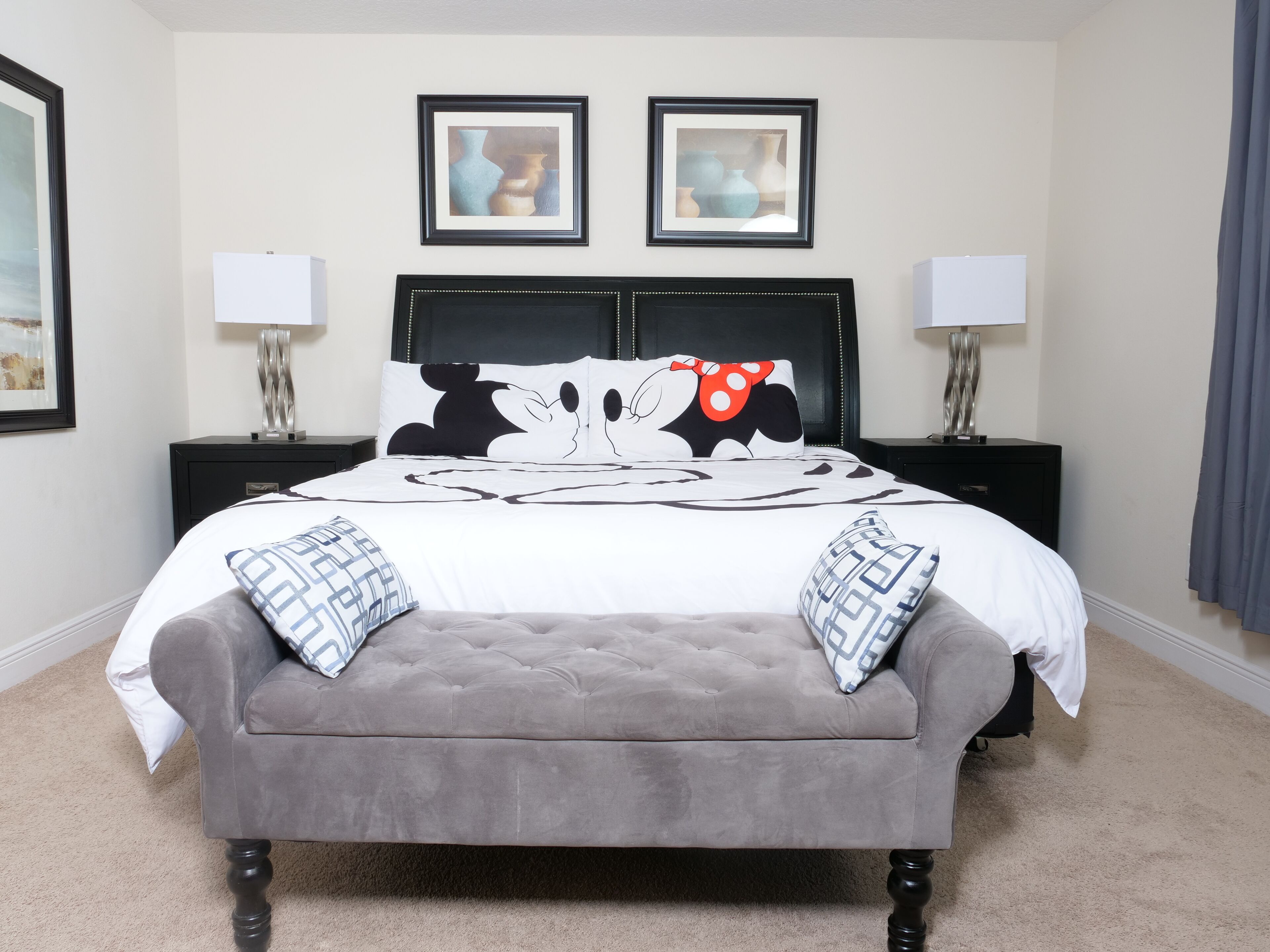 A neatly arranged bedroom with a tufted headboard, a grey upholstered bench, and two framed artworks above the bed.