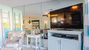 A living room with a television, a patterned armchair, a dining table, and a cabinet with electronic devices. - Beach Daze Sleeps 6 @ Gulf Highlands Dog Friendly (Panama City Beach)
