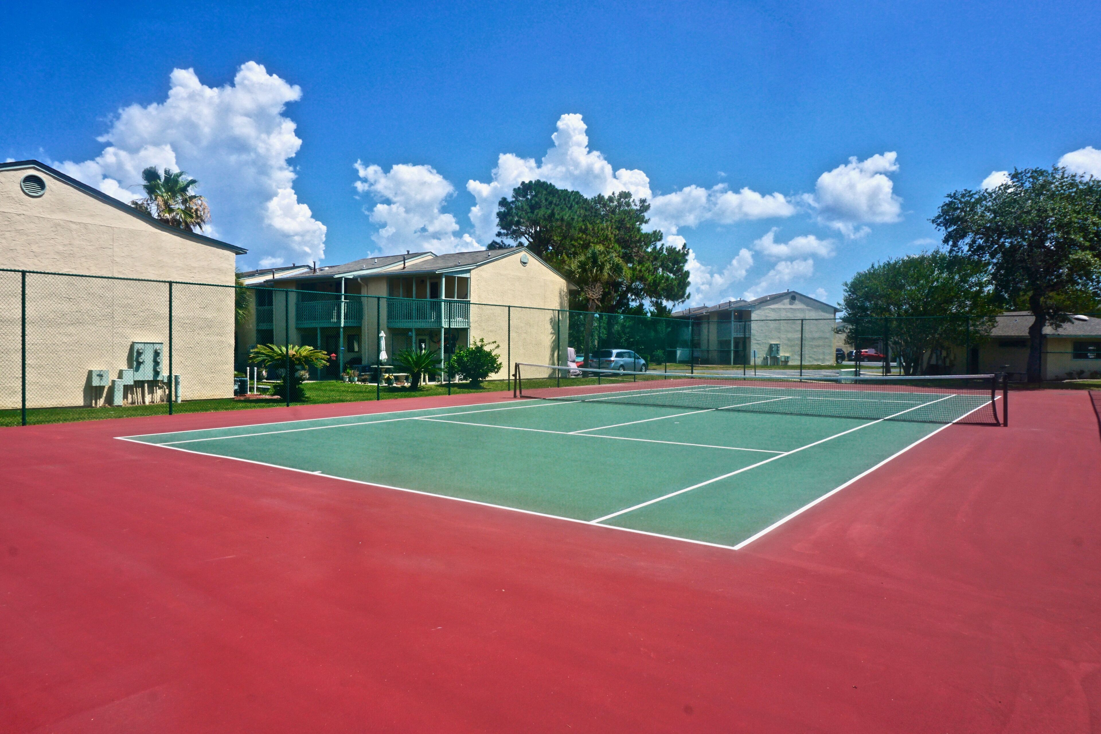 Una pista de tennis amb superfície vermella i verda, envoltada per una tanca, i al costat d'un edifici amb un balcó.