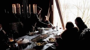 Une pièce faiblement éclairée, avec une table dressée pour un repas, des bougies et une vue sur les arbres dénudés à l’extérieur.