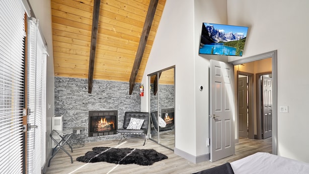 A modern room with a stone wall, wooden ceiling beams, a fireplace, a TV, and a bed.
