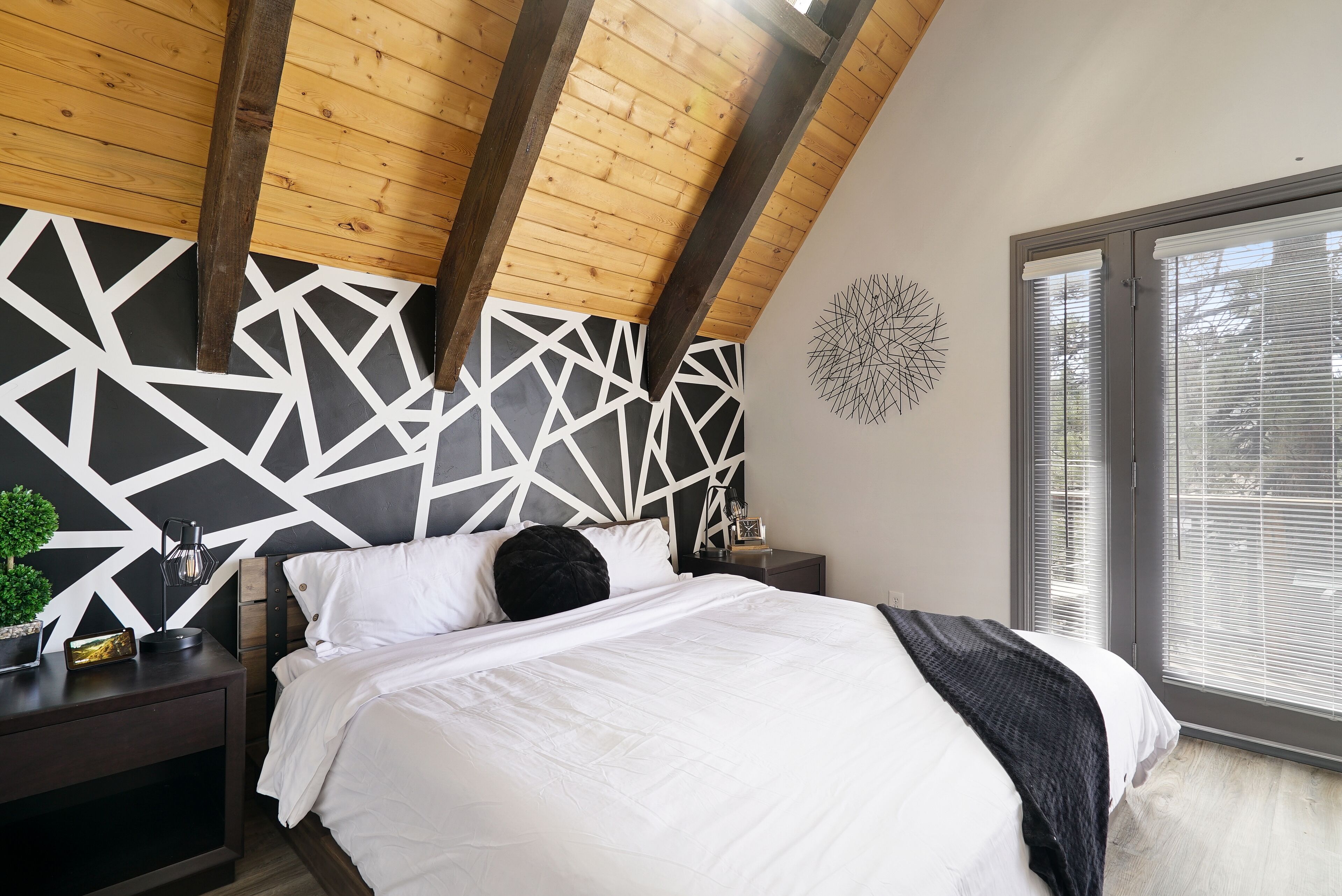 Un dormitorio con techo inclinado, cabecera de diseño geométrico, cama con ropa de cama blanca, mesita de noche con lámpara y ventana con persianas.